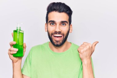 Sakallı genç yakışıklı adam aloe vera kremi şişesi giyiyor başparmağıyla yan tarafa bakıyor ve ağzı açık gülümsüyor. 