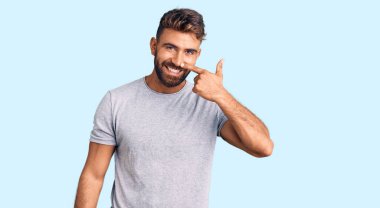 Latin asıllı genç bir adam, gündelik giysiler giyip el ve burnu işaret ediyor, neşeli bir şekilde gülümsüyor. güzellik konsepti 