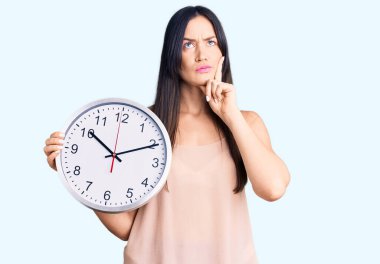 Genç, beyaz, güzel bir kadın. Büyük bir saat tutuyor. Ciddi bir yüz ifadesi var. 