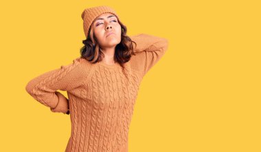 Genç, güzel, melez bir kadın yün kazak giyiyor ve kış şapkası takıyor. Boyun ağrısı çekiyor. Boynuna elinle dokunuyor. 