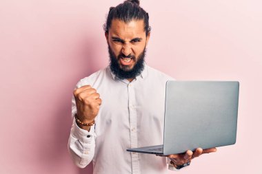 Genç Arap adam dizüstü bilgisayarını tutarken sinirlenmiş ve sinirlenmiş öfkeyle bağırıyor, öfkeyle deli gibi bağırıyor ve el kaldırıyor. 