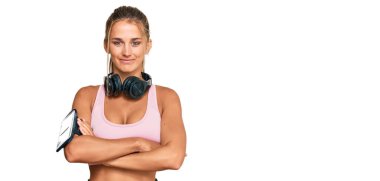 Spor kıyafetleri giyen ve kulaklık takan, kollarını kavuşturup kameraya gülümseyen genç sarışın kadın. pozitif kişi. 