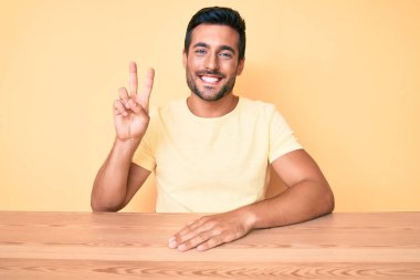 Gündelik kıyafetler giyen genç İspanyol adam masaya oturmuş gülümsüyor kameraya göz kırparak zafer işareti yapıyor. İki numara.. 