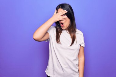 Genç, güzel, esmer, sıradan bir tişört giyen bir kadın mor arka planda dikiliyor. Şok olmuş yüzünü ve gözlerini eliyle kapatıyor, parmaklarından bakıyor.