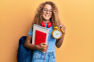 Güzel beyaz kız öğrenci çantası takıyor ve elinde çalar saatle mutlu ve havalı bir gülümsemeyle gülümsüyor. diş gösterme. 