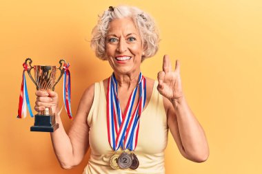 Kıdemli gri saçlı kadın elinde şampiyonluk kupası tutuyor. Madalya takıyor. Parmaklarıyla işaret veriyor, gülümsüyor. 