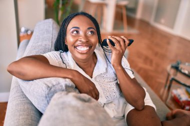 Genç Afrikalı Amerikalı kadın gülümseyerek evde akıllı telefon kullanarak mutlu mesut oturuyor.
