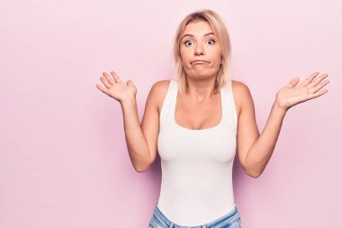 İzole edilmiş pembe arka planda duran sıradan tişört giyen genç ve güzel sarışın kadın. Hiçbir fikrim yok ve kuşkulu bir yüzüm var..