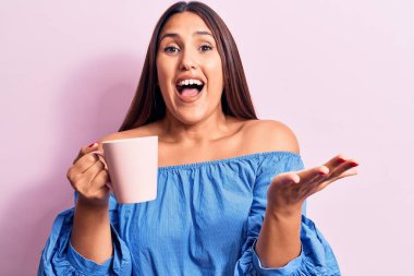 Genç, güzel esmer kadın bir kupa kahve içiyor başarıyı kutluyor mutlu bir gülümsemeyle ve kazanan yüz ifadesiyle elini kaldırarak. 