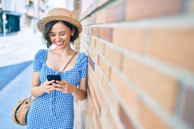 Genç ve güzel bir kız mutlu bir şekilde gülümsüyor. Akıllı telefon kullanıyor ve şehrin duvarına yaslanıyor.
