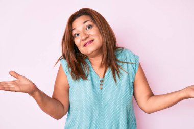 Orta yaşlı esmer, sıradan kıyafetler giyen, elleri ve kolları havada, kafası karışmış esmer bir kadın. şüphe kavramı. 