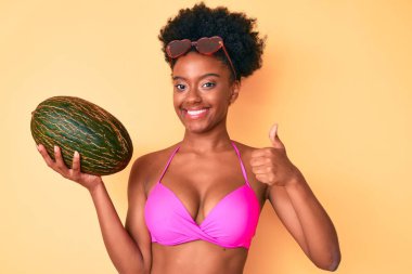 Genç Afro-Amerikalı kadın elinde kavun, mutlu ve pozitif gülümsüyor, başparmağını kaldırıyor ve onaylıyor. 