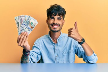 Masada oturmuş gülümseyen mutlu ve pozitif gülümseyen genç İspanyol adam başparmağıyla mükemmel ve onay işareti yapıyor. 