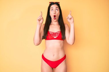 Bikini giyen güzel beyaz kadın şaşırmış ve şaşırmış bir şekilde yukarı bakıyor ve parmaklarıyla işaret ediyor ve kollarını kaldırıyor.. 