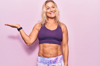 Pembe arka planda spor kıyafetleri giyen orta yaşlı, beyaz bir kadın. Gülümseyen, mutlu bir sunum yapan ve avuç içiyle kameraya bakan.. 