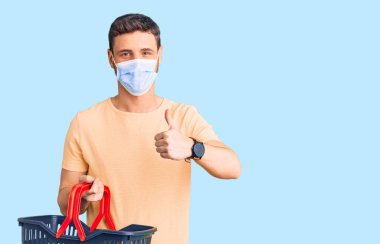 Alış veriş sepeti ve tıbbi maske takan yakışıklı genç adam mutlu ve olumlu gülümsüyor, başparmağıyla mükemmel ve onay işareti yapıyor. 
