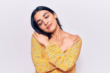 Genç, güzel, latin bir kadın gündelik kıyafetler giyip kendine sarılıyor, mutlu ve pozitif, kendinden emin gülümsüyor. Sevgi ve özgüven. 