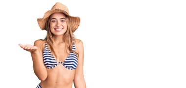 Bikini giyen, şapkalı, neşeli ve dostça kollarını açmış genç, güzel sarışın kadın. Hoş geldin, olumlu ve kendinden emin selamlar. 