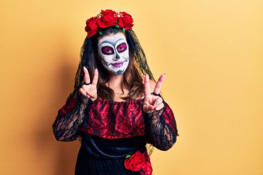 Sarı üzerine ölü kostümü giymiş genç bir kadın gülümsüyor ve parmaklarını zafer işareti yaparken kameraya bakıyor. İki numara.. 