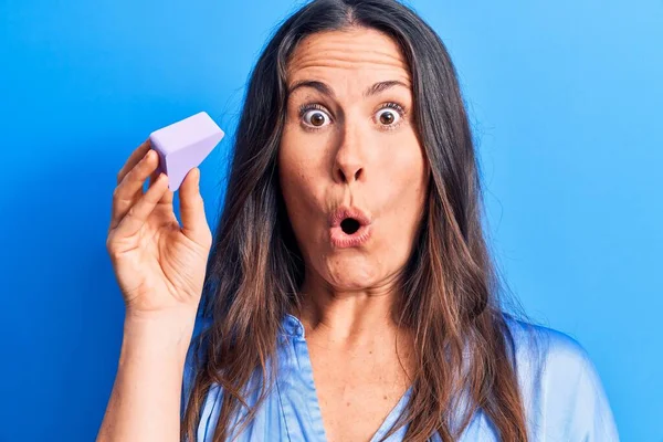 Genç, güzel, esmer bir kadın mavi arka planda sünger kullanarak makyaj yapıyor. Şaşırmak için ağzı açık ve korkmuş, inançsız bir yüz.