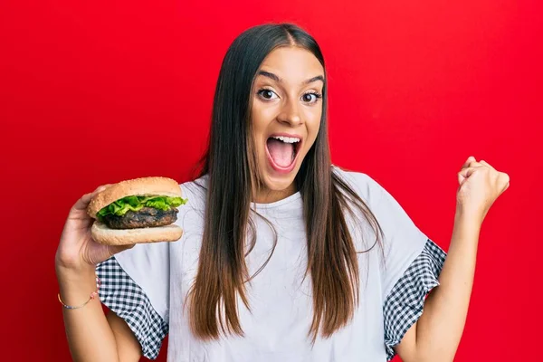 Young hispanic woman eating hamburger screaming proud, celebrating victory and success very excited with raised arms
