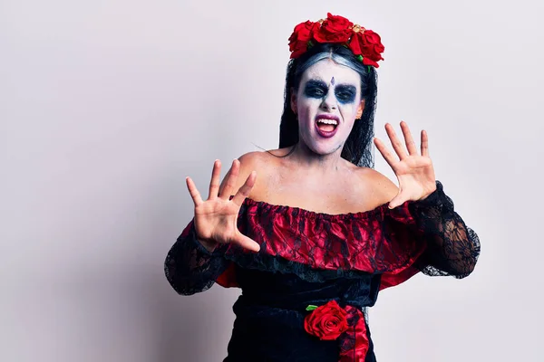 Young woman wearing mexican day of the dead makeup afraid and terrified with fear expression stop gesture with hands, shouting in shock. panic concept.