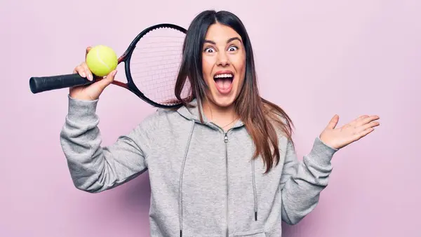 若い美しいスポーツ女性がラケットとボールを使用してテニスをプレイピンクの背景に幸せな笑顔で達成を祝い 手を上げて勝者の式 — ストック写真