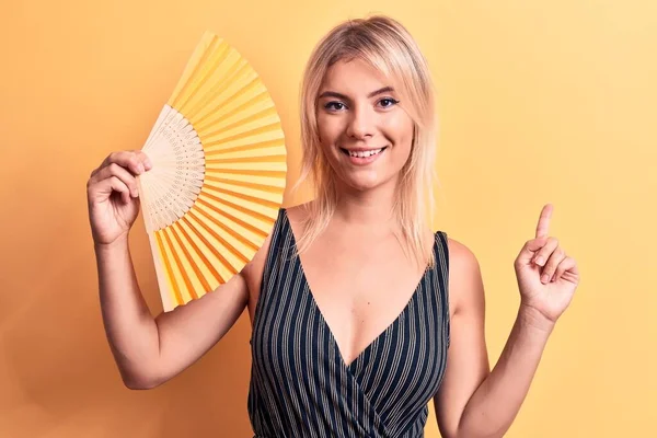Young Beautiful Blonde Woman Using Hand Fan Standing Isolated Yellow — Stock Photo, Image