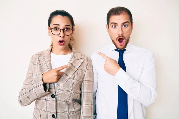 Beautiful couple wearing business clothes surprised pointing with finger to the side, open mouth amazed expression.