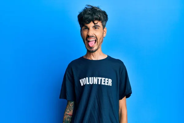 面白い表情で楽しそうに舌を突き出しているボランティアTシャツを着た若いヒスパニック系の男 感情の概念 — ストック写真