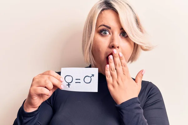 Blonde plus size woman asking for sex equality holding paper with gender equal message covering mouth with hand, shocked and afraid for mistake. Surprised expression