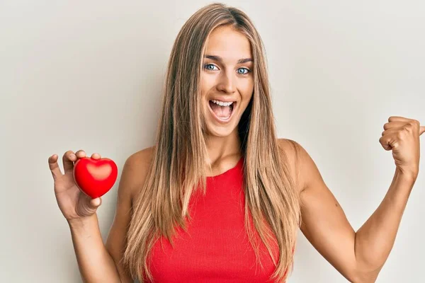 Joven Mujer Rubia Sosteniendo Corazón Señalando Pulgar Hacia Lado Sonriendo —  Fotos de Stock