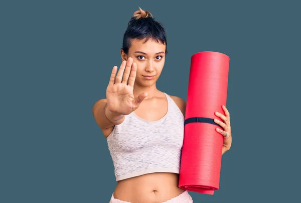 年轻女子张开手拿着瑜伽垫做停止手势 严肃而自信的表情 防守的姿势 — 图库照片