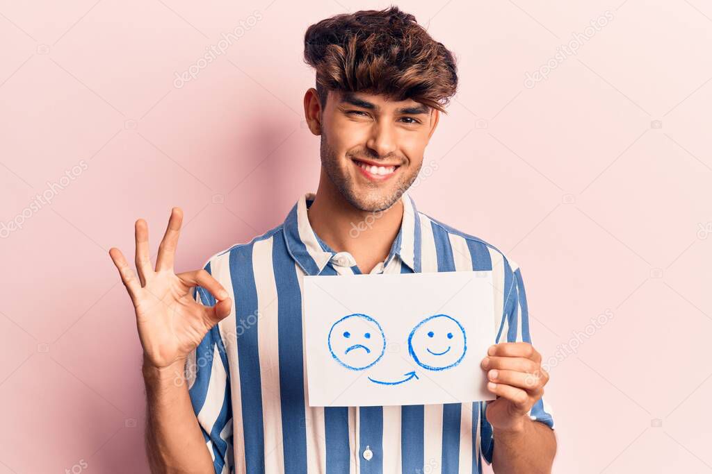 Young hispanic man holding sad to happy emotion paper doing ok sign with fingers, smiling friendly gesturing excellent symbol 