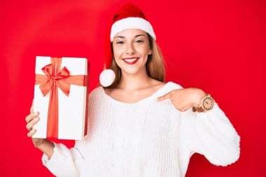 Genç esmer kadın Noel şapkası takıyor ve elinde bir hediye tutuyor. Gülümseyen mutlu ve gururlu birini işaret ediyor. 