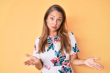 Orta yaşlı, çiçekli bir elbise giyen, elleri ve kolları havada, kafası karışık bir ifade takınan İspanyol bir kadın. şüphe kavramı. 