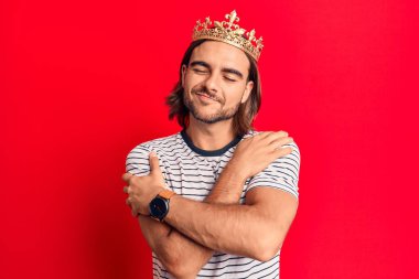 Prens tacı takan yakışıklı genç adam kendine sarılıyor mutlu ve pozitif, kendinden emin gülümsüyor. Sevgi ve özgüven. 