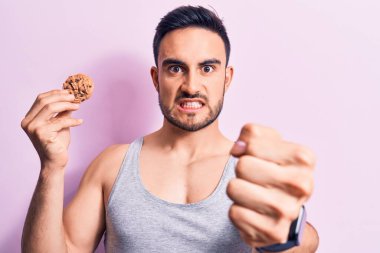 Sakallı, kolsuz tişört giyen, çikolatalı kurabiye yiyen sinirli ve sinirli genç bir adam öfkeyle bağırıyor ve el kaldırıyor.