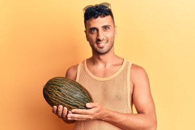 Karpuz tutan genç İspanyol adam pozitif ve mutlu görünüyor. Duruyor ve gülümsüyor. Kendine güvenen bir gülümsemeyle dişlerini gösteriyor. 