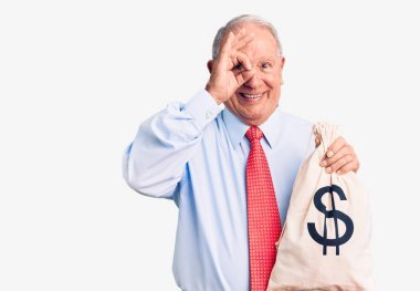 Üst düzey yakışıklı, gri saçlı, şık kıyafetler giyen, para çantası tutan mutlu mesut gülümseyen, el pençe divan duran bir adam. 