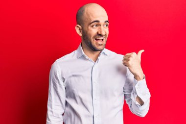 Zarif bir gömlek giymiş genç ve kel bir adam baş parmağını yana kaldırmış, ağzı açık gülümsüyor. 