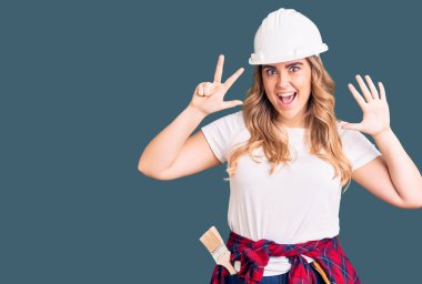 Güvenlik kaskı takan beyaz bir kadın sekiz numarayı gösterip işaret ederken kendinden emin ve mutlu gülümsüyor.. 