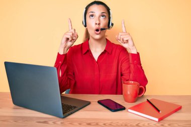 Çağrı merkezinde operatör kulaklığı takan genç esmer kadın şaşkın şaşkın yukarı bakıyor ve parmaklarıyla işaret ediyor ve kollarını kaldırıyordu.. 