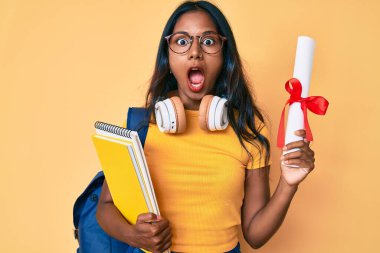 Sırt çantası taşıyan genç Hintli bir kız. Elinde diploma var. Şaşırmış ve şaşırmış bir ifade, korku ve heyecanlı bir yüz ifadesi var.. 