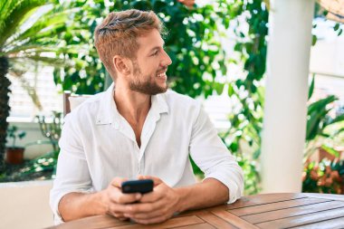 Evde terasta oturan yakışıklı beyaz adam mutlu bir şekilde akıllı telefon kullanıyor.