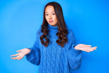 Kış süveteri giyen genç ve güzel bir Çinli kız. Kolları ve elleri havada, kafası karışmış bir ifade. şüphe kavramı. 