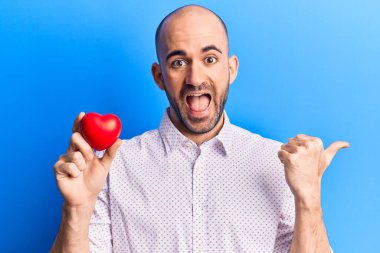 Genç, kel, yakışıklı, kalbi tutan, baş parmağını yana doğru kaldıran, ağzı açık gülümseyen bir adam. 