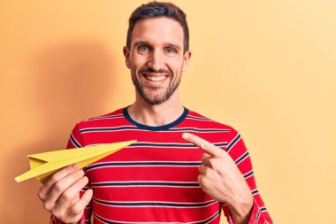 Sarı kağıt uçak tutan yakışıklı genç adam izole arka planda duruyor ve mutlu bir şekilde elini ve parmağını göstererek gülümsüyor.