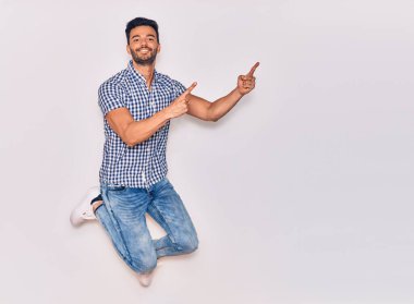 Genç, yakışıklı, sıradan kıyafetler giyen, mutlu gülümseyen bir İspanyol. Yüzünde tebessümle sıçrayıp, izole beyaz arka planda parmaklarını yana doğru gösteriyor.