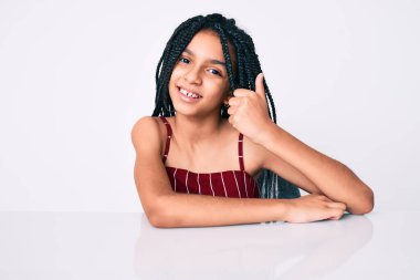 Saçları örgülü genç Afro-Amerikalı kız masada oturmuş, elleriyle mutlu bir jest yapıyor. Kameraya bakıp başarıyı gösteren ifade. 